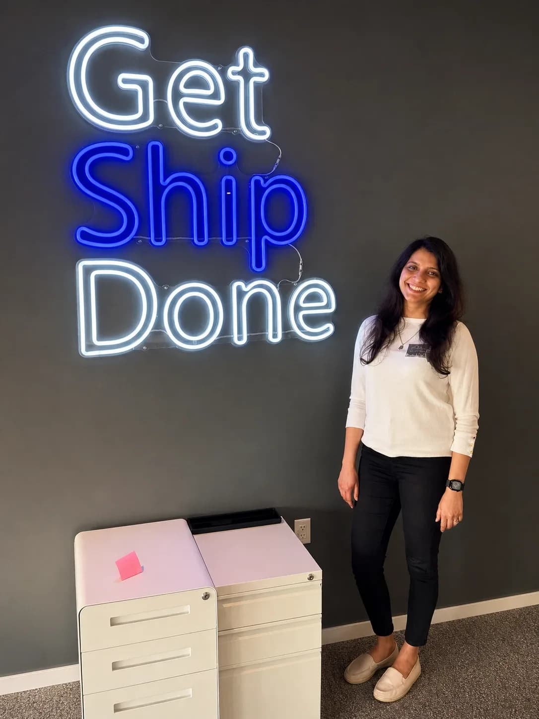 Sonali smiling beside a neon sign that reads Get Ship Done